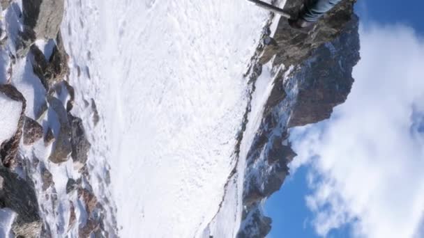 旅行者の垂直ビデオ映像は 風の強い冬の日に山でトレッキングを行います 雪のパン粉が地面に飛んでいる バックパックとトレッキングポールを持つ男 スローモーション — ストック動画