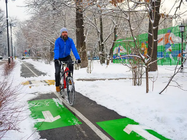 A bearded man rides a bicycle along a bike path in a park in winter. Active lifestyle in winter. Man in a blue jacket on a red bicycle