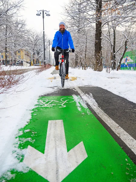 A smiling man rides a bicycle along a bike path in winter. Active lifestyle in winter. Bearded man in a blue jacket on a red bicycle