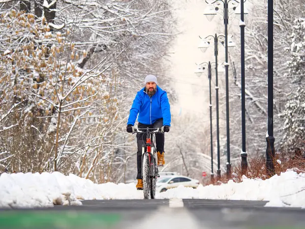 A bearded man rides a bicycle along a bike path in the city in winter. Active lifestyle. Guy in a blue jacket on a bicycle
