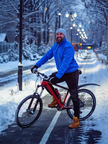 Bearded man with a bicycle in a winter city late in the evening. Active and healthy lifestyle in the city. Smiling man in a blue jacket with a red bicycle