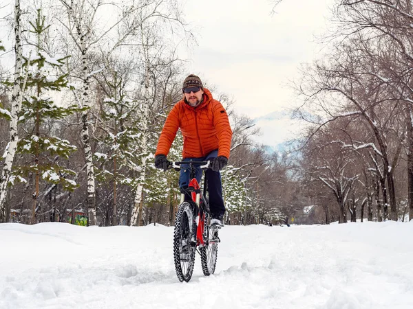 A guy rides a bicycle in a winter park. A cyclist in an orange jacket rides with difficulty along a snowy path. Eco-transport in the city