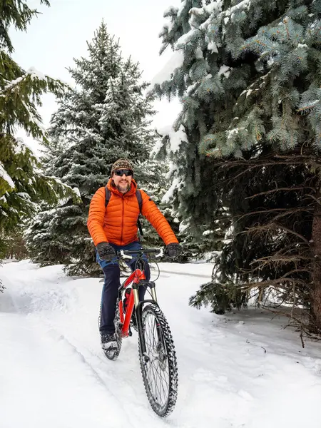 A guy rides a bicycle in a winter park. A cyclist in an orange jacket joyfully rides along a snowy path. Active lifestyle in winter