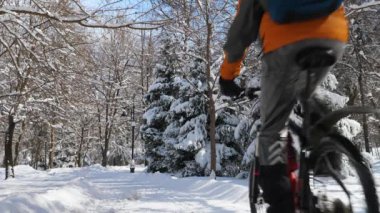 Güneşli bir kış gününde karla kaplı ağaçların arasında bisiklet süren bir adam. Kışın aktif bir yaşam tarzı. Turuncu ceketli, kasklı ve sırt çantalı bir adam. Arkadan bak. Yavaş çekim 2x
