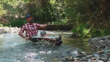 Sakallı bir adam nehirde bir turist sandalyesine oturur. Erkek gezgin, ağaçların gölgesinde bir dağ nehrinde tünemiş kahve içiyor.