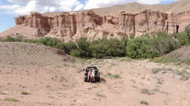 Sakallı erkek baba ve kızı kırmızı kayaların arasında muhteşem bir kanyonda bir kamyonetin bagajında oturuyorlar. Gezginler izleyicileri selamlamak için ellerini sallıyorlar.