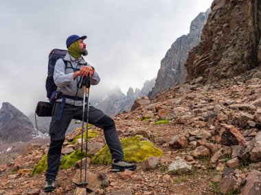 Dağlarda sakallı erkek gezgin. Sırt çantalı ve sırıkla dağlara tırmanan bir adam.
