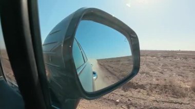 Issız bir araziden geçen bir arabanın dikiz aynasına bak. Araba tozlu bir toprak yolda gidiyor. Yavaş çekim 2x