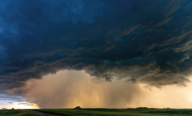 Şiddetli yağmur, ovalardaki yalnız bir evin üzerine karanlık bir süper hücre fırtınası bulutundan düşüyor. Güneş arkadan yağan yağmuru aydınlatıyor.