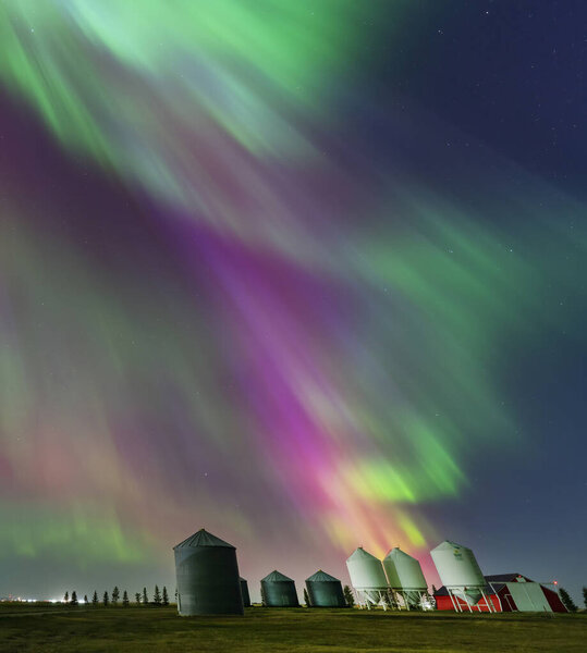 Colorful northern lights painting the night sky above grain silos and farm buildings