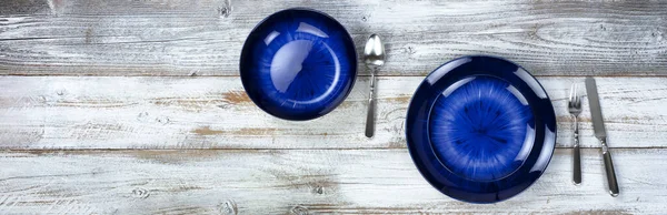 Basic dinner table setting of an empty main dark blue dish and bowl with cutlery on white wood in overhead or top view layout