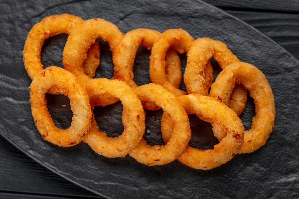 stock image Delicious golden battered, breaded and deep fried crispy onion rings served on round black stone tray on black wooden table, view from above, close-up