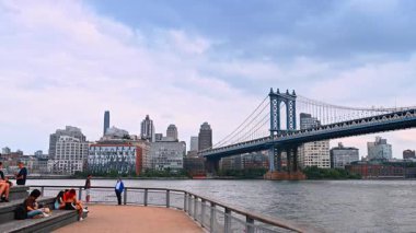 New York, ABD, 4 Temmuz 2025: İnsanlar New York, ABD 'deki East River' ın rıhtımında dinleniyor. Manhattan Köprüsü ve nehrin diğer yakası alçak açılı..