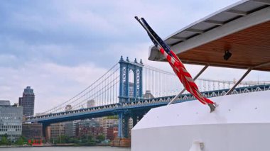 Nehirde dalgalanan Amerikan bayrağıyla dolu bir tekne. Bulutlu bir günde New York 'taki Manhattan Köprüsü' nün alçak açılı görüntüsü.