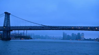 New York, ABD 'de alacakaranlıkta Williamsburg Köprüsü' nde ışıklar yanıyor. Nehir manzarasından bak. Arka planda sisli şehir silüeti.