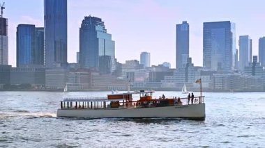 New York, ABD, 8 Temmuz 2025: Tek güverteli nehir teknesi gündüz vakti nehir kıyısına hareket ediyor. Modern New York, ABD 'nin puslu silueti.