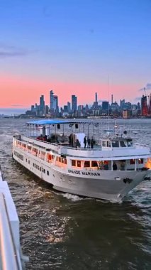 New York, ABD, 8 Temmuz 2025: Bir grup insan büyük yolcu teknesiyle seyahat ediyor. Günbatımında New York nehir turu. Pembe gökyüzünün arkaplanındaki şehir silüeti. Dikey video.