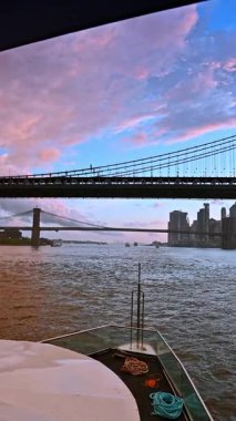 New York, ABD 'deki Manhattan Köprüsü' nün üzerindeki gökyüzünde sevimli pembe bulutlar. Doğu nehrinden geçen teknenin görüntüsü. Dikey video.