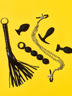 Black sex toys dildo, anal plugs and clamps over yellow paper background