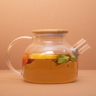 Portakal, böğürtlen ve biberiyeli meyve çayı, cam çaydanlıkta, ahududu tatlısı ve bej arka planda bir fincan ile servis edilir. Meyve çayı. Mevsimlik içki konsepti. Yapılandırma şablonu