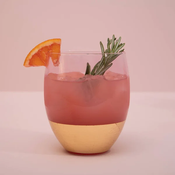 Elegant trendy, stylish pink gold cocktail glass with citrus fruit and rosemary on a pink background. Summer art food drink concept. Front view. Mockup template