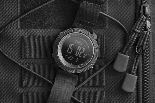 Tactical watch on backpack. Military field electronic watch in black steel case, having notched bezel, built-in compass, with nylon strap, lies on black tactical backpack, close-up.