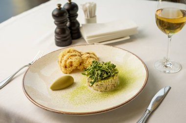 Beyaz bir tabakta mikro yeşilliklerle süslenmiş olivier salatası. Masada yulaf lapası ve bir bardak beyaz şarap ve çatal bıçakla.