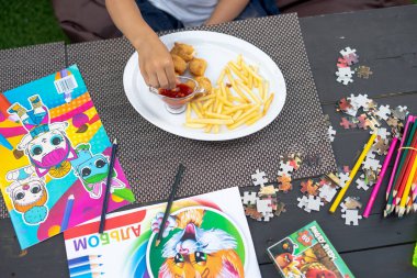 Çocuk patates kızartması, ketçap ve tavuklu nugget yiyor. Bulmacalar, kalemler ve boyama kitapları masanın üzerinde.