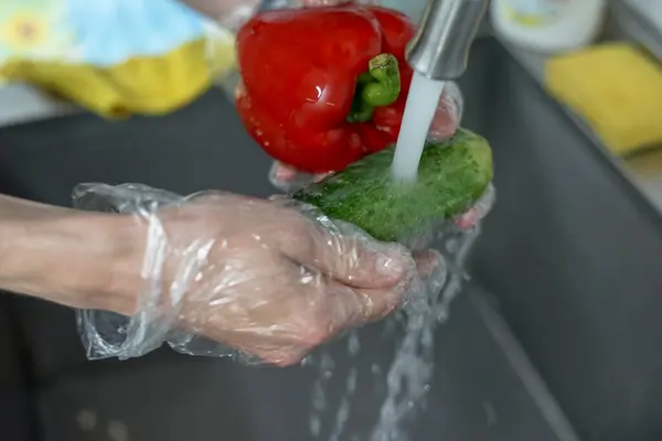 Kadın elleri koruyucu eldivenlerle mutfak lavabosunda biber ve salatalık yıkıyor.