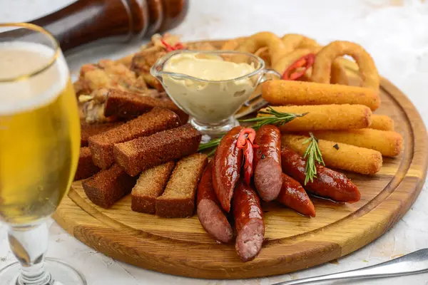 İçinde soğan halkaları, kanatlar, sosis ve bir bardak bira bulunan çeşitli bar atıştırmalıkları olan ahşap bir tabak..