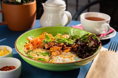 Tofu, kuşkonmaz, avokado parçaları, kırmızı marul ve kavrulmuş fıstık parçalarıyla servis edilen pirinç eriştesi. Yeşil bir kasede servis edilmiş. Sabah güneşiyle aydınlatılan bir kahvaltı seti.