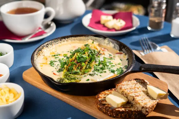 Çırpılmış yumurta, keçi peyniri ve sebzeli, keçi peyniri ve sebzeli, keçi buğday ekmeği, soğan soğanı, sarı peynir ve yeşillikle servis edilen dökme demir tavada. Sabah güneşiyle aydınlatılmış bir kahvaltı seti..