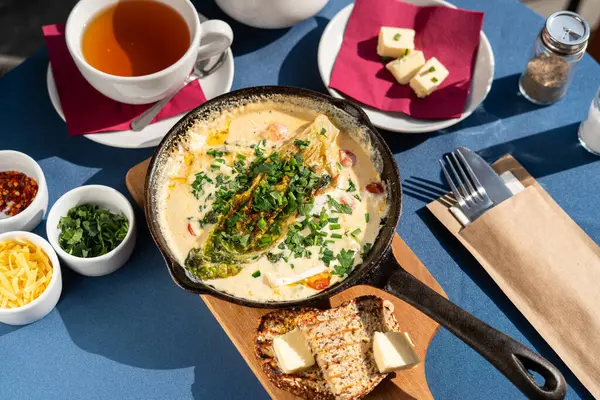 Çırpılmış yumurta, keçi peyniri ve sebzeli, keçi peyniri ve sebzeli, keçi buğday ekmeği, soğan soğanı, sarı peynir ve yeşillikle servis edilen dökme demir tavada. Sabah güneşiyle aydınlatılmış bir kahvaltı seti..