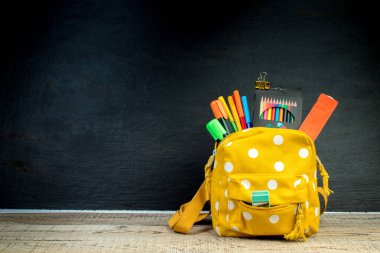 Masada beyaz benekli sarı sırt çantası ve farklı renkli kırtasiye kağıtları var. Siyah arka plan. Okula geri dön.