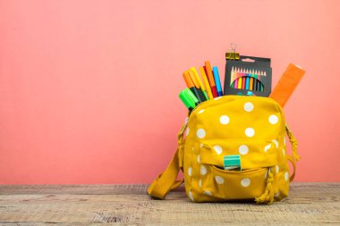 Masada beyaz benekli sarı sırt çantası ve farklı renkli kırtasiye kağıtları var. Pembe arka plan. Okula geri dön.