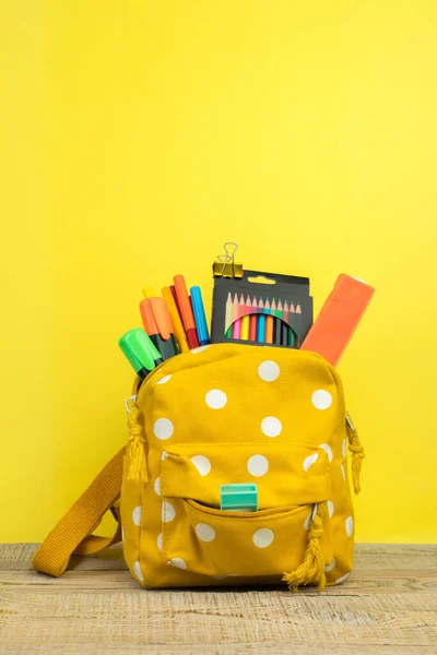 Backpack with different colorful stationery on table. Yellow background. Back to school