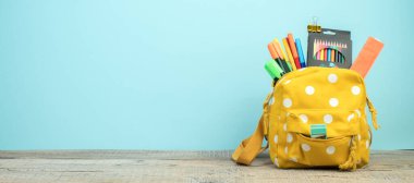 Masada beyaz benekli sarı sırt çantası ve farklı renkli kırtasiye kağıtları var. Mavi arka plan. Okula dönelim. Pankart tasarımı