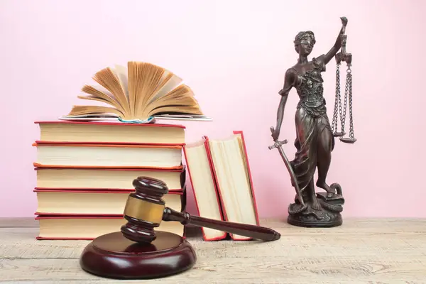 Law concept - Open law book, Judge's gavel, scales, Themis statue on table in a courtroom or law enforcement office. Wooden table, pink background.
