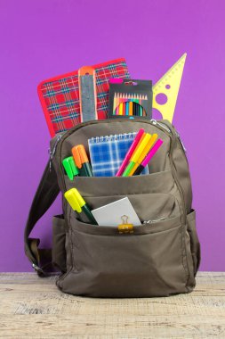 Sırt çantasında farklı renkte kırtasiye malzemeleri var. Mor arka plan. Okula geri dön.