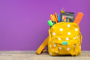 Sırt çantasında farklı renkte kırtasiye malzemeleri var. Mor arka plan. Okula geri dön..