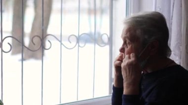 Elderly woman, grandmother, puts on a mask and looks out the window.