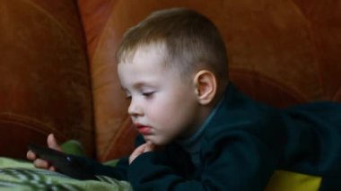 Küçük çocuk kanepede otururken telefonda oynuyor..