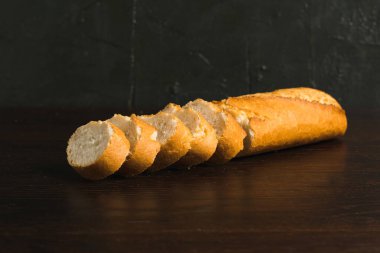 French baguette with appetizing crust cut into pieces on wooden table close-up, dark wooden background with copy space.
