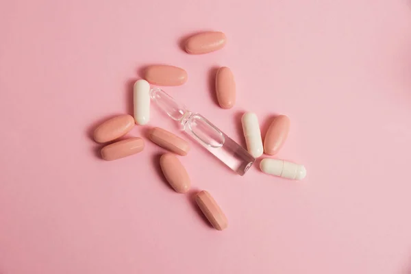 Pink pills, white medical capsules and ampoule on pink paper background with copy space. The concept of taking vitamins in tablet form and in injections.