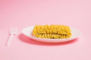 Beyaz tabakta, pembe arka planda plastik çatalla hazır şehriye. Modern dünyada fast food.