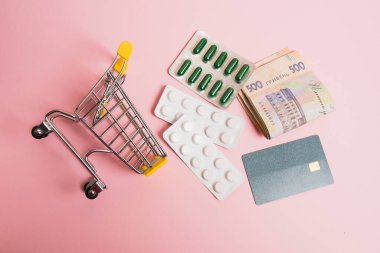 Flatlay, alışveriş arabası, banka kartı ve pembe bir arka planda su toplayan haplar..