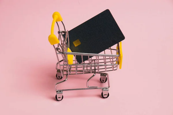 Bank card in the shopping cart on a pink background. Concept of inflation, rising prices for food and medicine