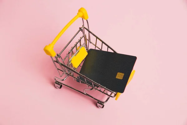 Bank card in the shopping cart on a pink background. Concept of inflation, rising prices for food and medicine