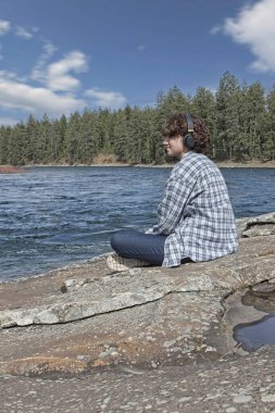 Post Falls, Idaho 'da Spokane nehrinin kenarında oturan genç bir kadın güneşli bir günde dinleniyor ve kulaklıklarıyla müzik dinliyor..