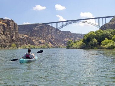 Genç bir çocuk, Idaho, Twin Falls 'daki Snake River' da güzel bir günde Perrine Köprüsü 'ne doğru kanoyla kürek çekiyor..